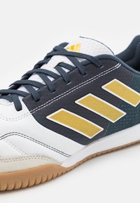 Scarpa da ginnastica bianca con suola blu scuro e in gomma, caratterizzata da tre strisce gialle texturizzate e dettagli grigi. I lacci sono di colore blu scuro.