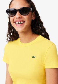 Geel geribbeld T-shirt met korte mouwen en een klein groen krokodillenlogo. Model draagt zwarte cat-eye zonnebril en glimlacht.