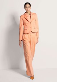 Perzikkleurige blazer met twee voorzakken en bijpassende getailleerde broek. Glanzende oranje hoge hakken maken de outfit compleet voor een gecoördineerde look.