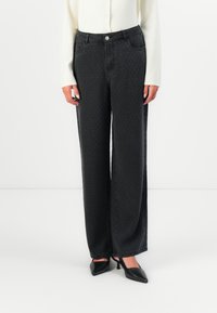 Woman wearing high-waist wide-leg black textured pants, white long-sleeve top, and black pointed-toe heels on a white background.