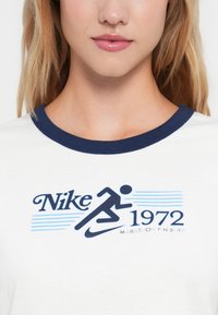 Balts t-krekls ar tumši zilu ribinātu apkakli. Uz tā ir zila grafiskais teksts "Nike 1972" un skrējēja attēls, ar "MARATHON FINISHER" zemāk.