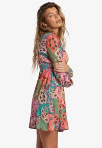 Frau mit welligem Haar, die ein buntes Blumenkleid mit langen Ärmeln trägt und über ihre Schulter zu einem schlichten Hintergrund schaut.