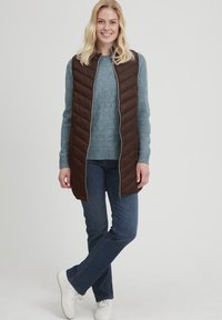 Brun quiltet vest med fuld lynlås, båret over en lyseblå struktureret sweater, parret med blå jeans og hvide sneakers. Neutral baggrund.