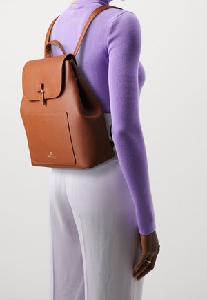 Femme portant un haut moulant à manches longues couleur lavande et un pantalon blanc, avec un sac à dos structuré en cuir marron sur le dos.