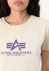 Bežinė marškinėlių su apykakle tipo T, pasižyminti didele violetine spausdinta logotipu su stilizuotu „A“ ir juostelėmis. Minkšta medvilnė, trumpomis rankovėmis.
