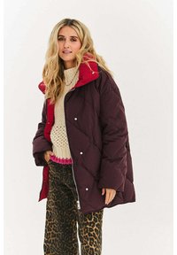 Bordo puchowa kurtka z czerwonymi akcentami, pikowana w diamenty, z zatrzaskami. Noszona na kremowym dzianinowym swetrze i spodniach w cętki lamparta.