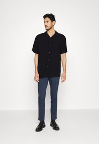 Chemise noire à manches courtes avec boutons, en tissu léger, associée à un pantalon bleu marine coupe slim et des chaussures habillées noires.