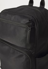 Sac à dos en nylon noir avec plusieurs poches zippées, une texture lisse et deux poignées supérieures pour le transport.