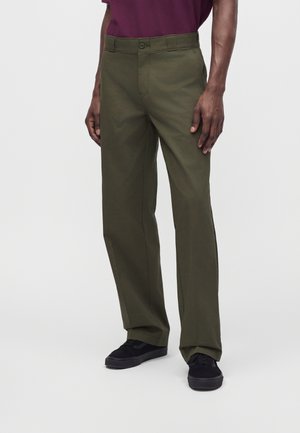 Uomo che indossa pantaloni verde oliva a gamba dritta abbinati a sneakers nere e una camicia viola, in piedi davanti a uno sfondo bianco semplice.