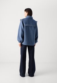 Plava flis jakna s traper yoke-om i čipkastim obrubom, s visokom kragnom i oversized rukavima, uparenim s tamnim trapericama.