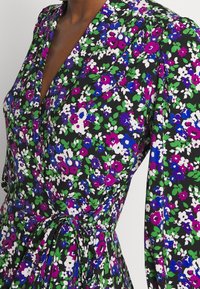 Blommig omlottklänning i svart tyg, med ett livfullt mönster av lila, blå och gröna blommor. Knytning i midjan och långa ärmar.