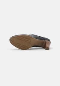 Zapato de cuero negro con un tacón de madera delgado. La suela presenta un patrón ondulado para ofrecer agarre y soporte. Diseño simple y funcional.