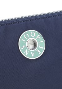 Námořnická látková taštička se zipem, vybavená kulatým stříbrným kovovým prvkem s vygravírovaným textem a světle zeleným pozadím uprostřed.