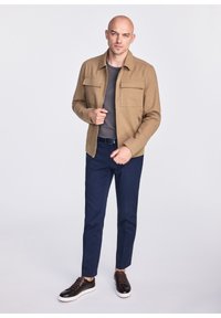 Beżowa, lekka kurtka z dwoma kieszeniami na piersi, szary t-shirt z okrągłym dekoltem, granatowe spodnie i ciemnobrązowe trampki z teksturowanymi sznurówkami.