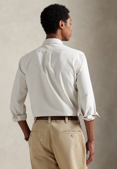 Vyras dėvi šviesią dryžuotą ilgomis rankovėmis marškinius su susuktiems rankogaliais ir smėlio spalvos kelnes su ruda diržu, žiūrint iš nugaros.