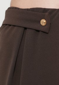 Rjava krilca z gubami iz teksturirane tkanine, z ravnim pasom in zlate barve gumbom spredaj.