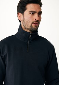 Czarny pulower na zamek wykonany z gładkiego materiału. Posiada wysoki kołnierz i płaskie szwy. Dopasowany krój, miękka struktura.