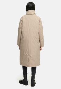 Beige lang frakke med høj krave, glat tekstur, sidelommer og lige snit, parret med sorte ankelstøvler og mørke jeans.