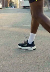 Schwarzer Sportschuh mit weißer Sohle, verfügt über ein schlankes Design, Schnürsenkel und ein markantes Logo; getragen mit einer weißen Socke, auf einer Straßenoberfläche.