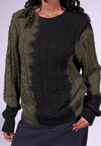 Personne portant un pull en maille torsadée vert foncé et noir à manches longues, associé à une jupe sombre, main posée sur la hanche.