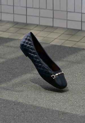 Chaussure plate matelassée noire avec détail de boucle en métal doré, présentée sur un trottoir gris près d’un mur carrelé.