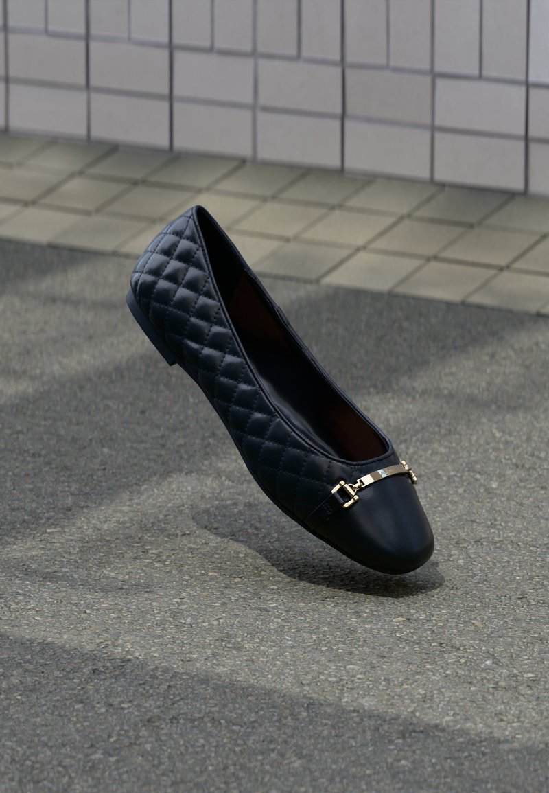 Schwarzer gesteppter flacher Schuh mit goldfarbenem Metallverschluss, gezeigt auf grauem Pflaster vor einem gekachelten Wandhintergrund.