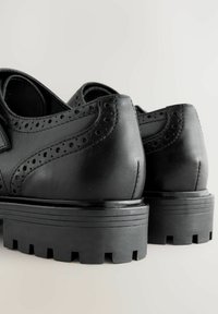 Mustat nahkakengät, joissa on brogue-yksityiskohtia, pyöreä kärki ja paksu kumipohja, jossa on uria ja pyöreitä aukkoja kantapään lähellä.