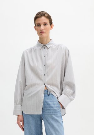 Frau trägt ein hellgraues, vertikal gestreiftes Hemd mit übergroßen Ärmeln und hellblaue Jeans, steht vor einem weißen Hintergrund.