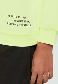 Neon gele sweatshirt met zwarte tekst die luidt: "REALITEIT IS MAAR EEN SUGGESTIE, IK DROOM ANDERS." Met manchetmouwen en een zachte textuur.
