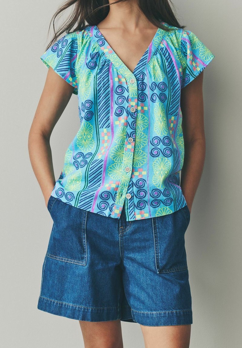 Femme portant un chemisier coloré à manches courtes avec boutons, comportant des motifs bleus, verts et violets, associé à un short en denim bleu avec poches.