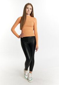 Oranje geribbelde lange mouwen top gecombineerd met zwarte neplederen leggings en meerkleurige sneakers. Steil haar valt over de schouders.