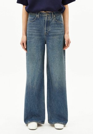 Person trägt eine hoch taillierte, weit geschnittene blaue Jeans mit Knopf und Reißverschluss vorne, kombiniert mit weißen Schuhen und einem dunklen Oberteil.