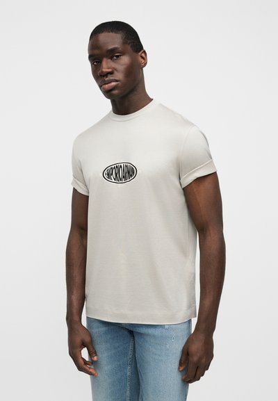 Ανοιχτό μπεζ κοντομάνικη μπλούζα από μαλακό βαμβάκι με στρογγυλή λαιμόκοψη και μαύρο οβάλ λογότυπο τυπωμένο μπροστά. Εφαρμοστή γραμμή.