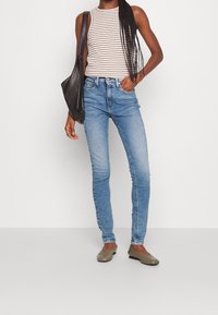 Pantalones vaqueros ajustados en denim azul claro, con cintura alta, cinco bolsillos y un ligero desgaste. Combinados con una camiseta de tirantes a rayas y zapatos planos.