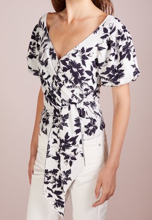 Weiße Wickelbluse mit marineblauem Blumenmuster, kurzen Puffärmeln, tiefem V-Ausschnitt und langem Frontband, kombiniert mit weißen Hosen.