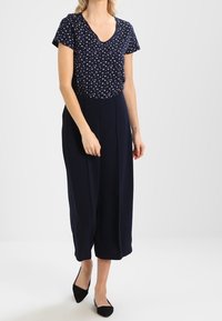 Mono azul marino de manga corta, escote en V, patrón floral y pantalones de pierna ancha. Acabado con tacones negros. Textura de tela suave.