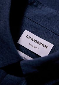 Collo a camicia in cotone blu navy con un'etichetta che dice "LINDBERGH RELAXED FIT." Il testo è nero su uno sfondo bianco. Tessuto morbido e con una texture.