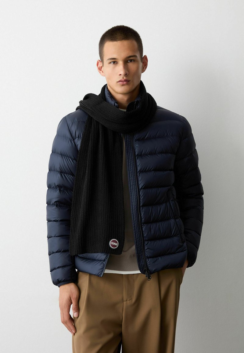 Giubbotto imbottito navy con trama trapuntata, sciarpa in maglia nera e pantaloni marroni; presenta una toppa con logo sulla sciarpa per il branding.
