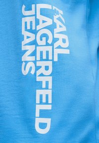 Φωτεινό μπλε ύφασμα με το λευκό κάθετο κείμενο "KARL LAGERFELD JEANS" τυπωμένο καθαρά πάνω του.