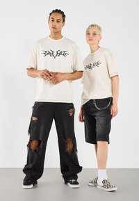 Svetle béžové oversized trička s černými grafickými designy, v kombinaci s černými roztrhanými džíny a džínovými šortkami. Doplňky zahrnují prsteny a řetízky.