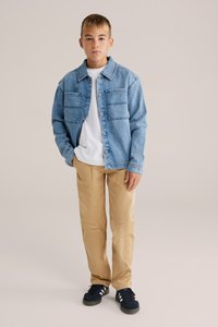 Šviesiai mėlynas džinsinis švarkas su dviem krūtinės kišenėmis, dėvimas ant baltos marškinėlių, derinamas su chaki spalvos kelnėmis ir juodais sportbačiais.