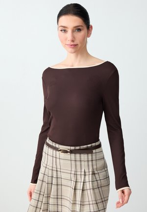 Donna con capelli scuri raccolti, che indossa una maglia a maniche lunghe marrone scuro e una gonna beige a quadri con una cintura marrone, in piedi davanti a uno sfondo semplice.