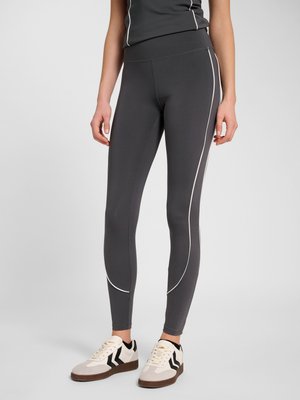 Persoon draagt donkergrijze sportlegging met witte biezen, zwart-witte sneakers, staand tegen een lichtgrijze achtergrond.