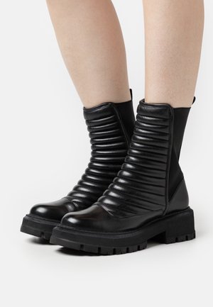 Botins pretos até ao tornozelo com um acabamento em couro brilhante, detalhes canelados no cabedal e solas de borracha grossa para maior aderência.