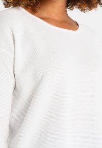 Jersey blanco texturizado con un ajuste holgado, que presenta un cuello redondo ancho. La tela tiene un patrón tejido sutil y un acabado suave.