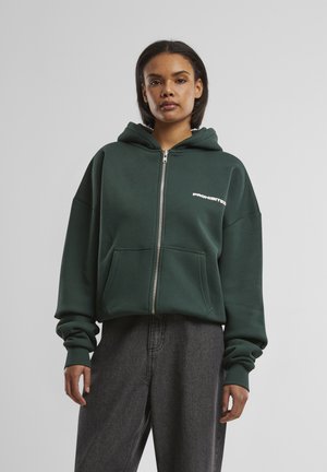 Grüne Zip-Hoodie aus weichem Material, mit einer Fronttasche und dem Wort "VERBOTEN" in Weiß auf der oberen linken Seite.