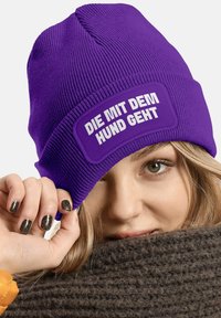 Purple knit beanie with a textured finish and a rectangular patch displaying white text. Soft material visible around the edges.