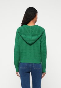 Zöld kötött pulóver kapucnival, texturált mintázattal, bordás mandzsettákkal és aljjal. A hátsó nézetben egy szűk szabás látható.