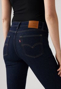 Jeans de color azul oscuro con un diseño ajustado, que presentan un parche trasero de cuero marrón y costuras doradas. Incluyen dos bolsillos traseros y una etiqueta roja.