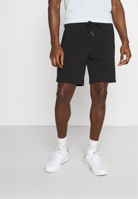 Shorts de algodón negros con cintura ajustable, corte a la rodilla y bolsillos laterales, combinados con zapatillas blancas y calcetines hasta el tobillo.